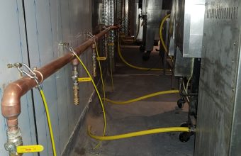 Copper gas manifold with isolation valves and yellow flexible hoses behind commercial catering appliances